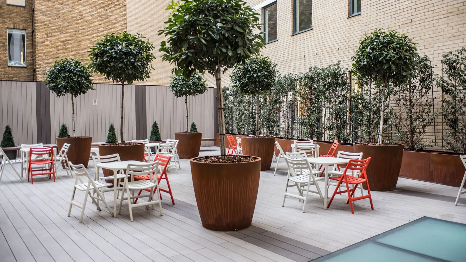 Clipped standard trees in tall corten steel planters on a decked terrace with glass rooflights, between pale London brick walls