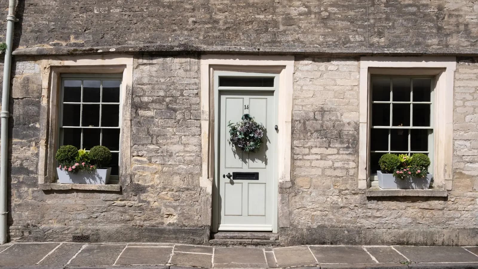 A pale green front door set in a Cotswold stone cottage, with clipped box balls in grey window troughs either side