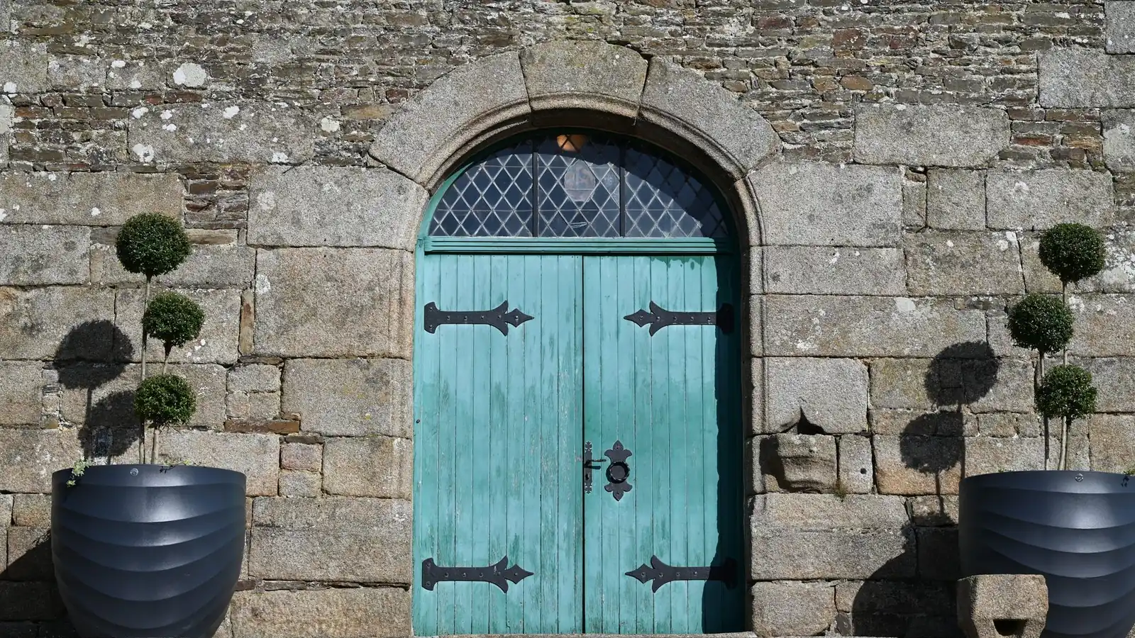 Two large charcoal ribbed planters holding tiered box topiary either side of a turquoise double door in an old granite stone wall